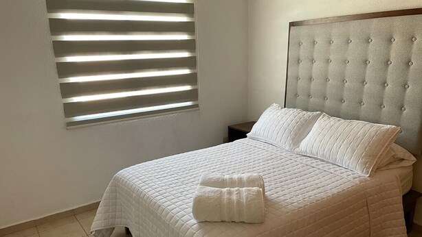 Ein ordentlich bezogenes Bett mit gesteppter weißer Bettdecke und Kissen, einem gepolsterten Kopfteil und einem Fenster mit horizontalen Rollläden.