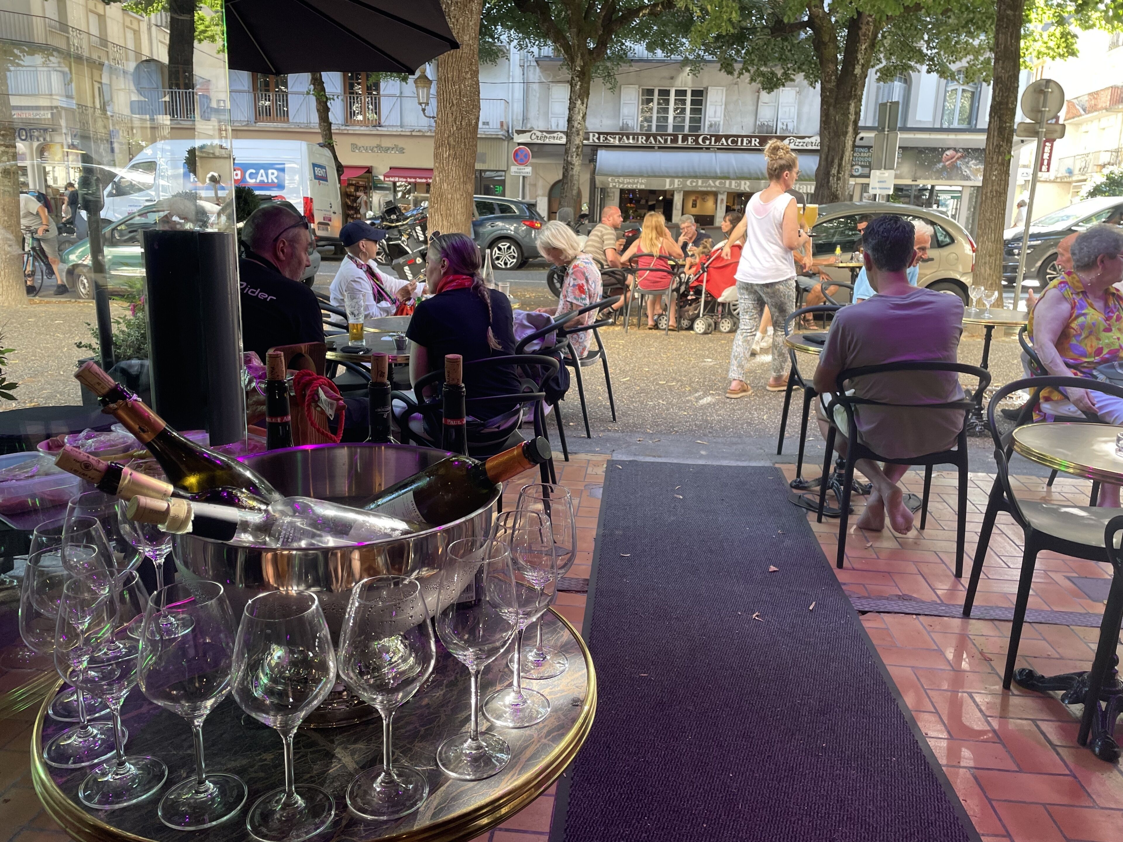 Un espacio exterior para comer con mesas y sillas, un gran cubo metálico con botellas de vino y copas, y vista a la calle con peatones y coches estacionados.