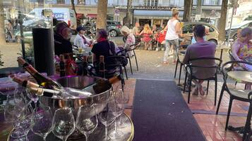 Un espacio exterior para comer con mesas y sillas, un gran cubo metálico con botellas de vino y copas, y vista a la calle con peatones y coches estacionados.