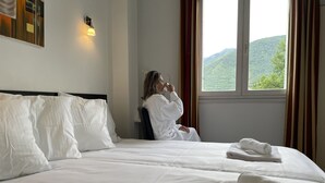 Habitación de hotel con una cama grande, ropa de cama blanca, vistas a colinas verdes y una ventana con cortinas.