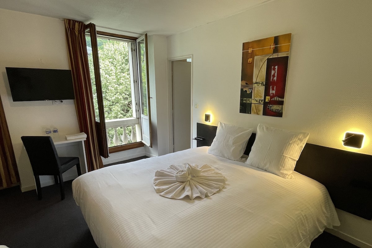 Habitación de hotel con una cama grande, un escritorio, una silla, un televisor montado en la pared y un ventanal con cortinas.