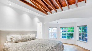 A bedroom with a large bed, wooden ceiling beams, and large windows with a view of greenery.