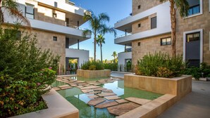 A modern building with a courtyard featuring a reflective water surface, surrounded by planters and palm trees. - Gorgeous apartment in Orihuela (Orihuela)