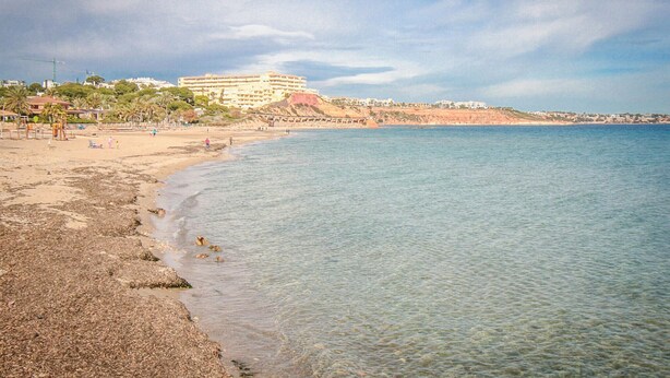 A beach with clear water, a sandy shore, and a large building with multiple balconies in the background. - Gorgeous apartment in Orihuela (Orihuela)