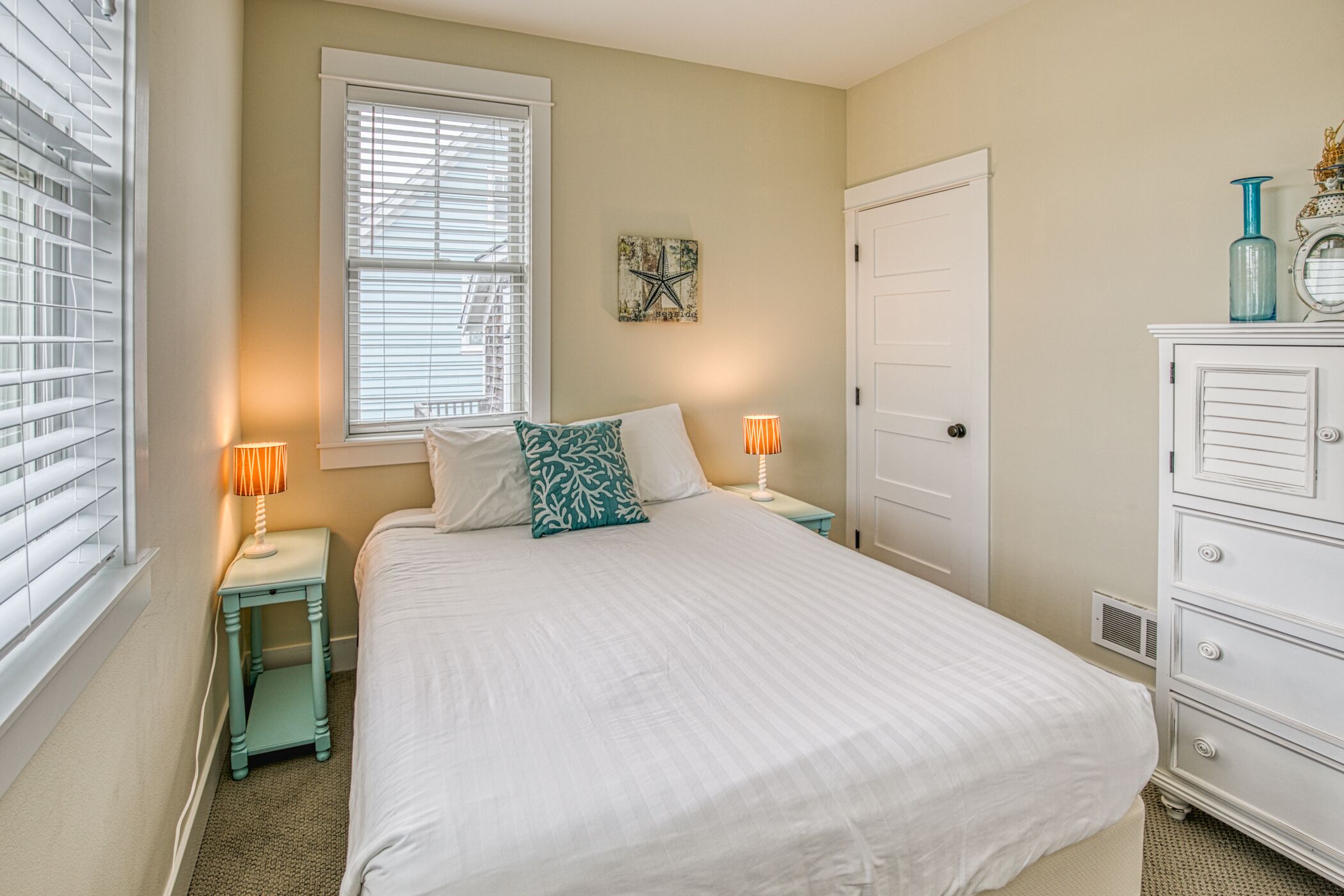 A bedroom with a large bed, two bedside tables with lamps, a dresser, and a window with blinds.