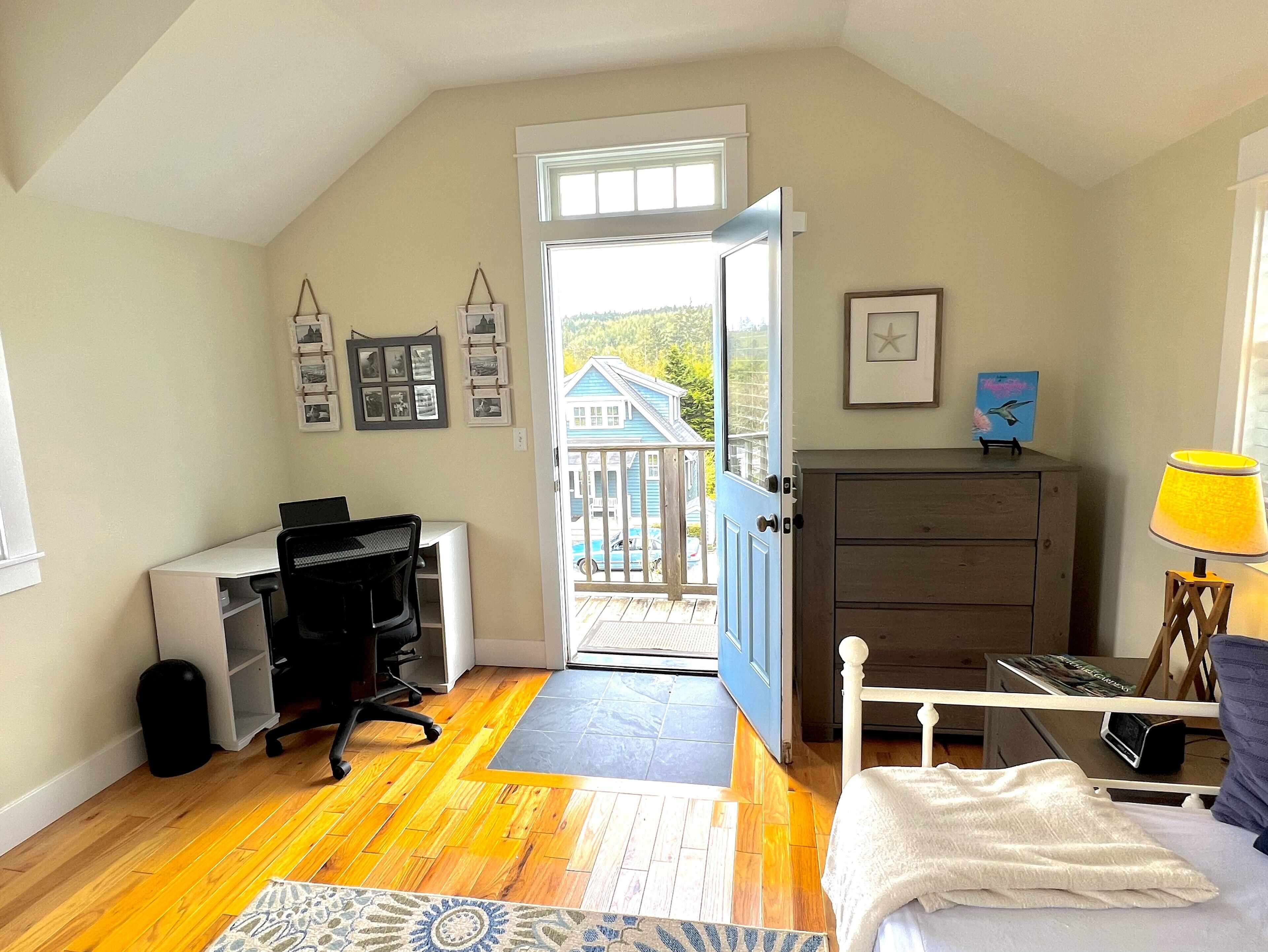 Un dormitorio con escritorio, cómoda, cama y vista a una casa azul a través de una puerta abierta.