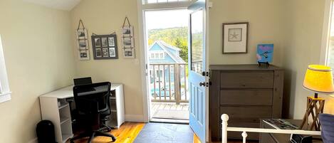Un dormitorio con escritorio, cómoda, cama y vista a una casa azul a través de una puerta abierta.