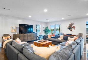 A modern living room with a large grey sectional sofa, a wooden coffee table, a flat-screen TV, and a dining area with a table and chairs.