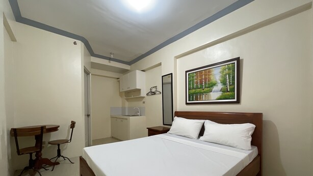 A bedroom with a bed, a desk, a chair, a small kitchenette, and a painting on the wall. - Lawton Residences powered by Cocotel (Taguig)