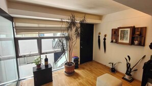Een moderne woonkamer met een groot raam, houten vloeren, een potplant, een zwart kastje en een lijst met een foto aan de muur.