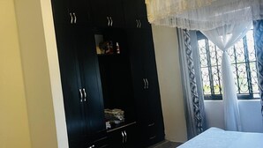 A bedroom with a large wardrobe, a bed, and a window with curtains. - House - Sleeps 6 - Parking - Garden (Kampala)