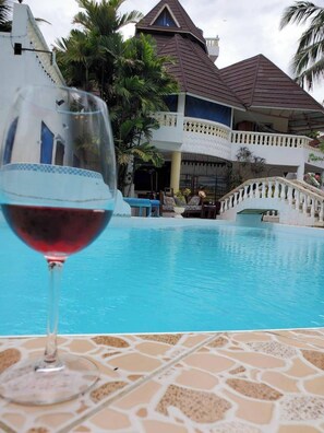 Une piscine, avec un verre de vin rouge au premier plan, un bâtiment blanc avec un toit brun, et des palmiers en arrière-plan.