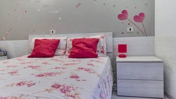 Una camera da letto con un letto, due cuscini rossi, un comodino con una lampada rossa e una parete decorata con cuori e la scritta 'Love'.