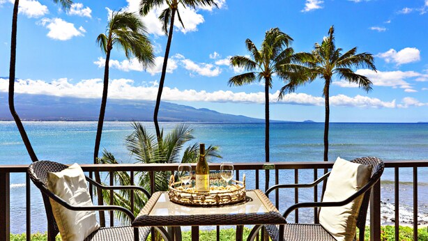A balcony with wicker chairs and a table set with a bottle and glasses, overlooking a beach with palm trees and the ocean.