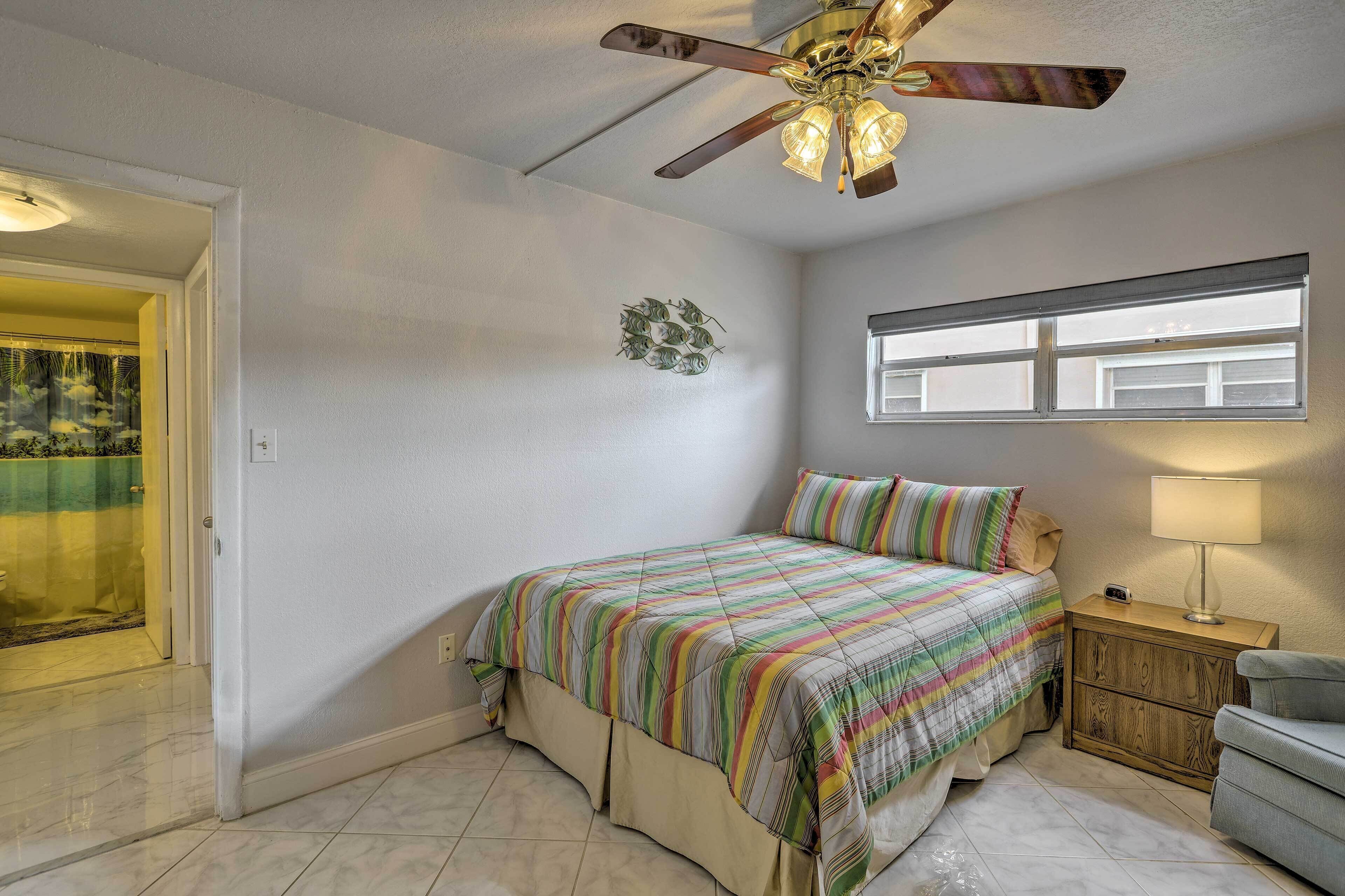 Une chambre à coucher comprenant un lit, une table de chevet, une chaise, un ventilateur de plafond et une fenêtre.