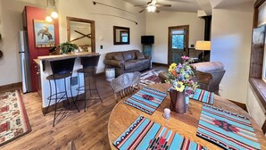 A modern kitchen with a bar area, a dining table with colorful placemats, and a living room with a sofa and a TV.