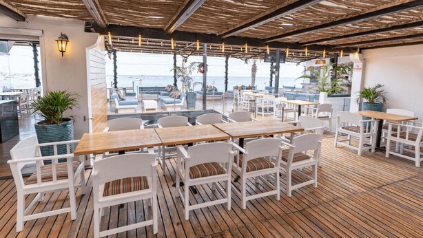 Un área de estar al aire libre con mesas de madera y sillas blancas, con vistas a un puerto deportivo lleno de barcos.