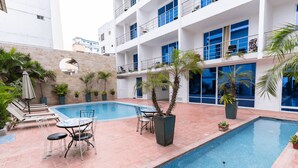 Zona de piscina de hotel con piscina rectangular, sillas de descanso y una mesa con sillas.