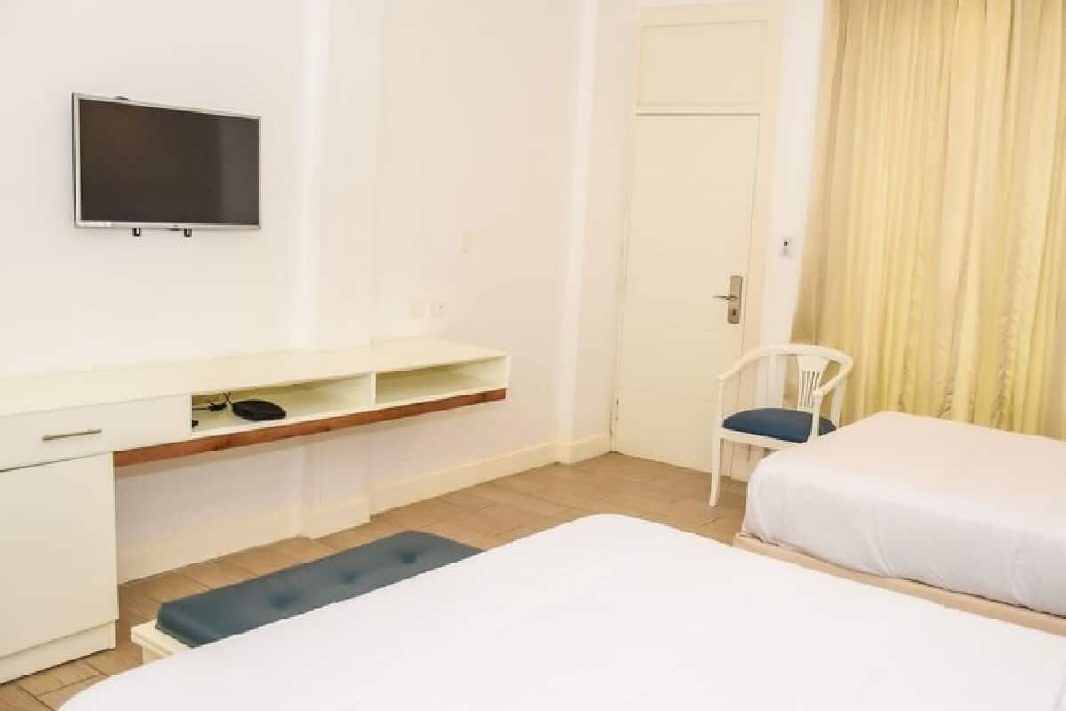 A hotel room with a bed, a desk with a chair, a TV mounted on the wall, and a door.