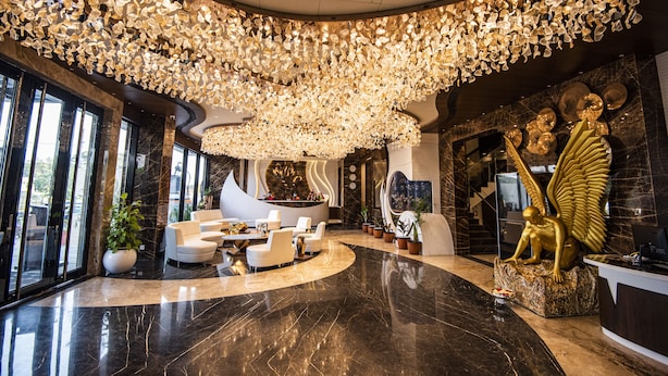 Un vestíbulo lujoso con un techo alto decorado con candelabros de cristal, zonas de estar elegantes y un mostrador de recepción con una estatua dorada.