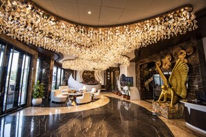 Un vestíbulo lujoso con un techo alto decorado con candelabros de cristal, zonas de estar elegantes y un mostrador de recepción con una estatua dorada.