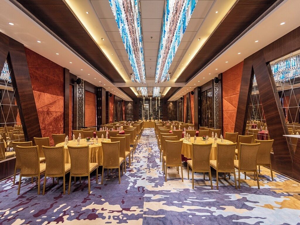A banquet hall with round tables and chairs, a patterned carpet, and a modern chandelier.