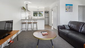 A modern living room with a leather sofa, a round coffee table, and a TV on a wooden stand. A kitchen with white cabinets and a stainless steel refrigerator is visible in the background. - Renovated Takapuna 2BR Apt - Parking (Auckland)