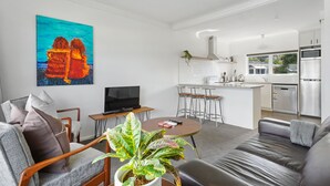 A modern living room with a sofa, a coffee table, and a TV. There is a kitchen area with a dining table and chairs. - Renovated Takapuna 2BR Apt - Parking (Auckland)