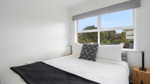 A bedroom with a large bed, white bedding, a black throw blanket, and a window with a view of trees and buildings. - Renovated Takapuna 2BR Apt - Parking (Auckland)