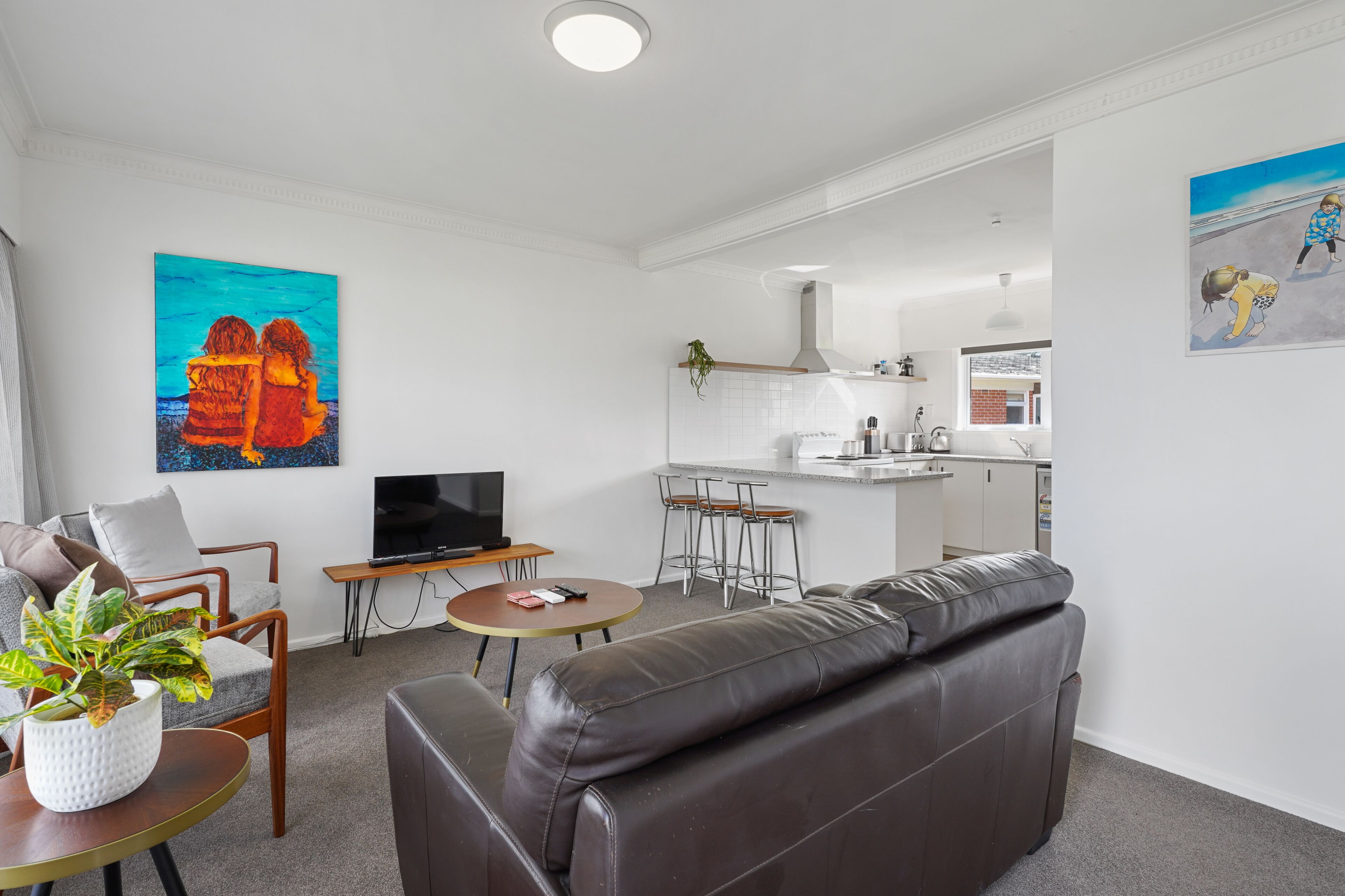 A modern living room with a leather sofa, a small round table, and a TV. There is a kitchen area with a bar stool and a plant on the counter.