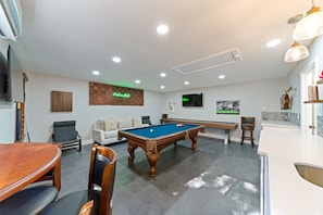 A pool table in a room with a white ceiling, recessed lighting, a wooden table, and a white sofa.