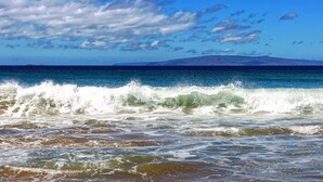 Una playa con olas y una cadena montañosa distante bajo un cielo parcialmente nublado.
