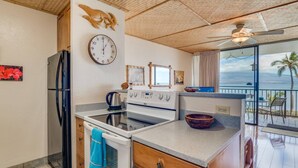 Una cocina con estufa, horno y refrigerador. Hay un reloj en la pared y una ventana con vista al océano.