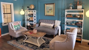 A cozy living room with blue walls, a grey sofa, a white armchair, a coffee table, and a bookshelf. - Mountainside Inn (Fort Davis)