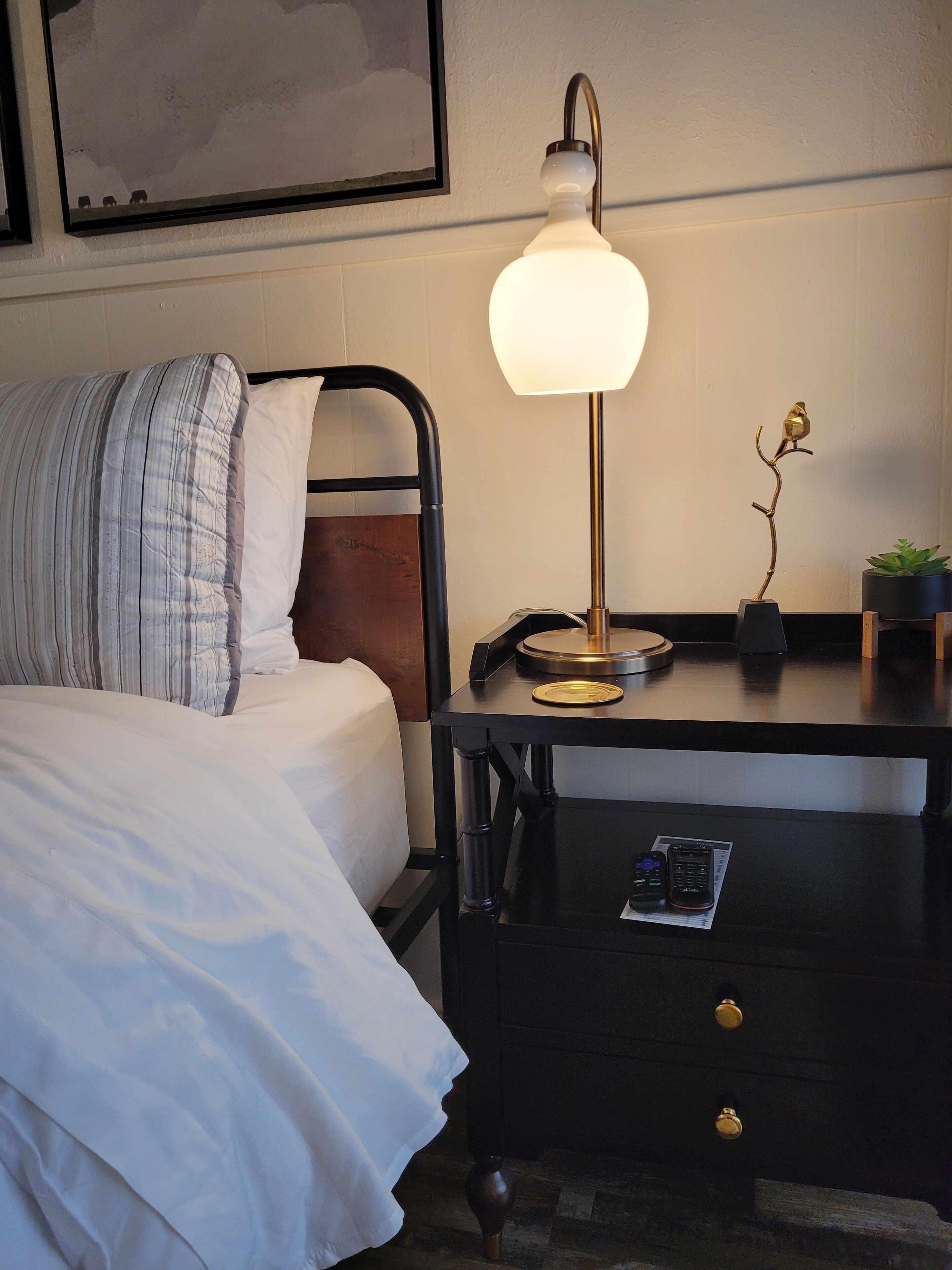 A neatly made bed with white linens, a black metal headboard, a bedside table with a lamp, and a small potted plant.