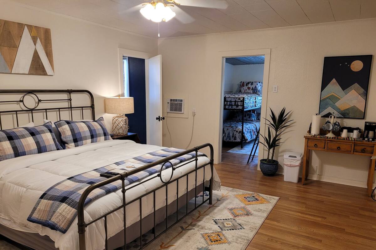 A bedroom with a bed, bedside table, lamp, and a view into a bedroom with a bunk bed.