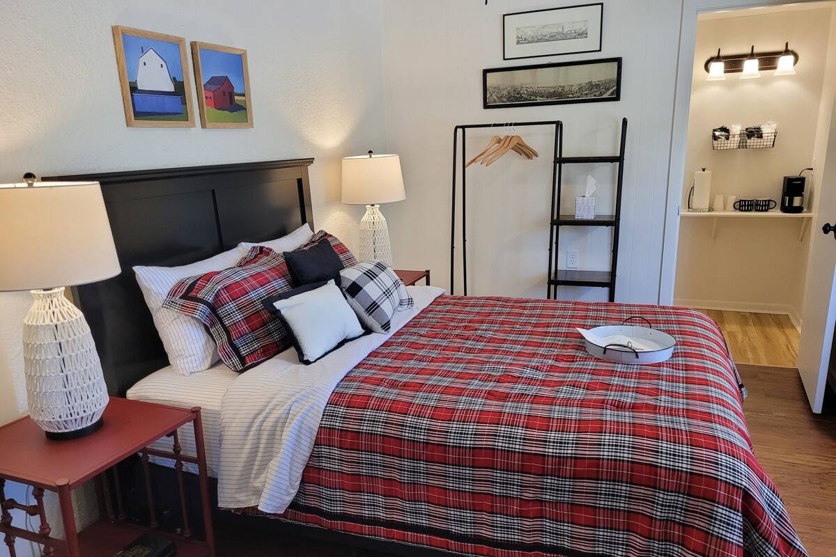 A neatly made bed with a red and black checkered bedspread, two bedside tables with lamps, a wooden nightstand, and a wall-mounted shelf with hanging clothes.