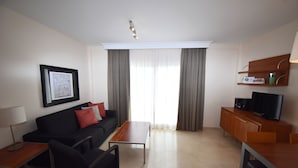Una habitación de hotel moderna con un sofá negro, una mesa de centro de madera, un televisor sobre un mueble de madera y un cuadro enmarcado en la pared.