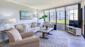 Un salon moderne avec un canapé, des fauteuils, une table basse et un téléviseur. Un tableau orne le mur, une plante est posée à proximité, et de grandes fenêtres offrent une vue sur les bâtiments environnants.