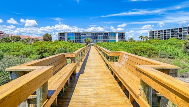 Une promenade en bois mène à un bâtiment moderne à plusieurs étages, doté de balcons et entouré d’une végétation luxuriante.