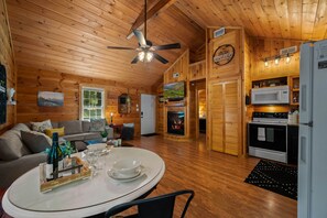 A cozy cabin interior with a wooden ceiling, a dining area with a table set for two, a fireplace, a kitchen with modern appliances, and a comfortable living room with a sofa and decorative pillows.