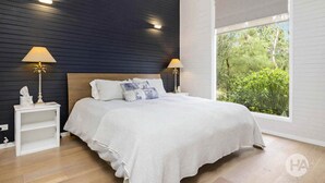 A bedroom with a dark wooden headboard, white bedding, two bedside lamps, and a large window with a view of greenery. - Coolart Studios: Studio Two (Balnarring)