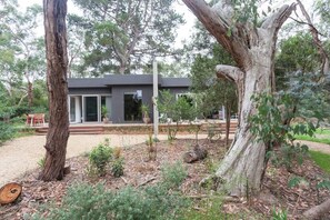 Een modern huis met een grote boom op de voorgrond, omgeven door een tuin met diverse planten en een houten bankje.