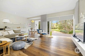 Een moderne woonkamer met een bank, salontafel en televisie. Er is een eethoek met stoelen en een tafel. De kamer heeft houten vloeren en grote ramen.