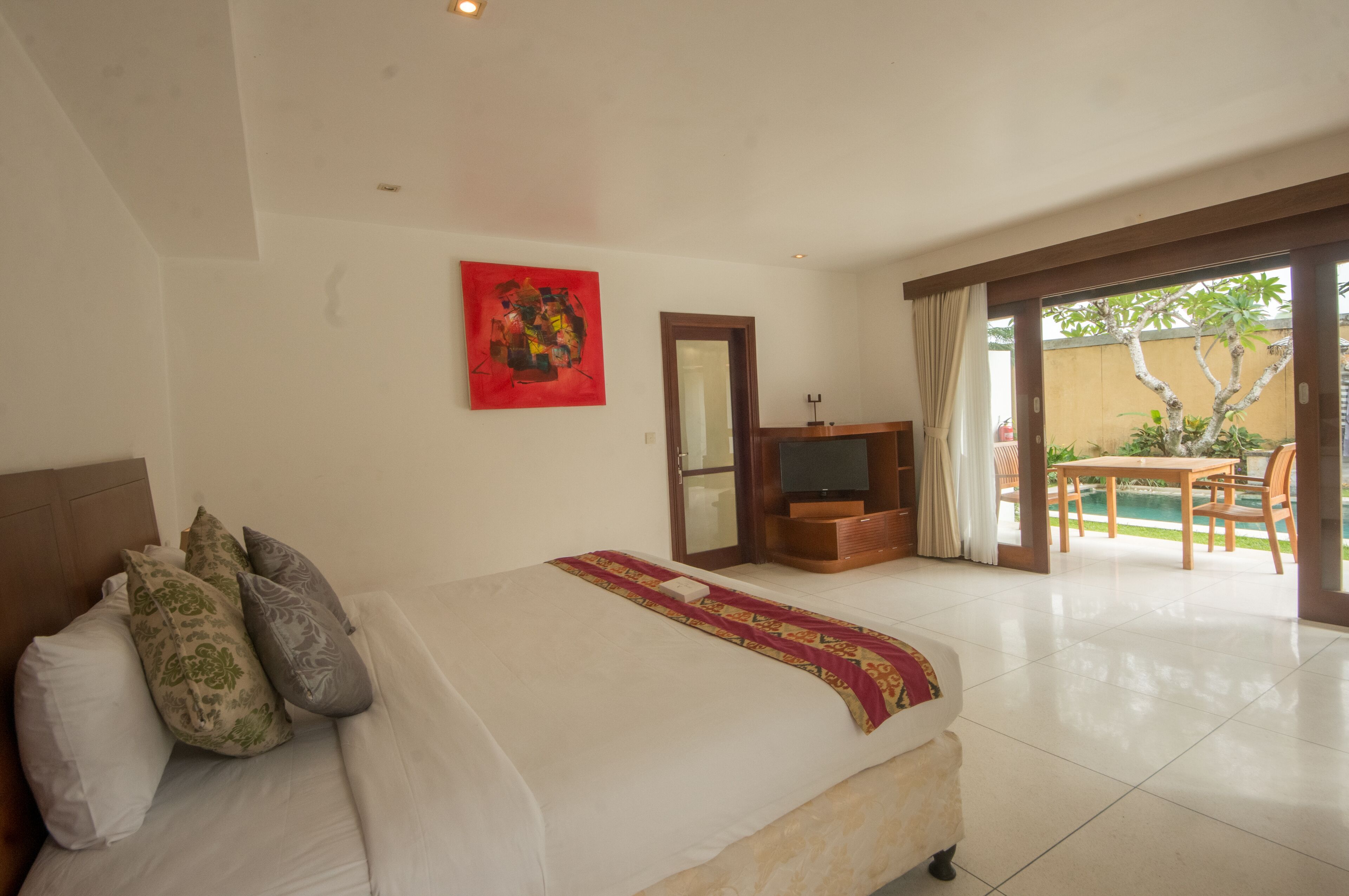 A hotel room with a bed, a TV, a wooden cabinet, and a sliding glass door leading to an outdoor area with a table and chairs.