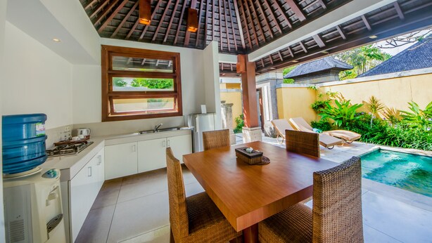 A modern kitchen with white cabinets, a dining area with a wooden table and wicker chairs, and a view of a swimming pool. - Nomad Hub Canggu-Bali (Kerobokan)