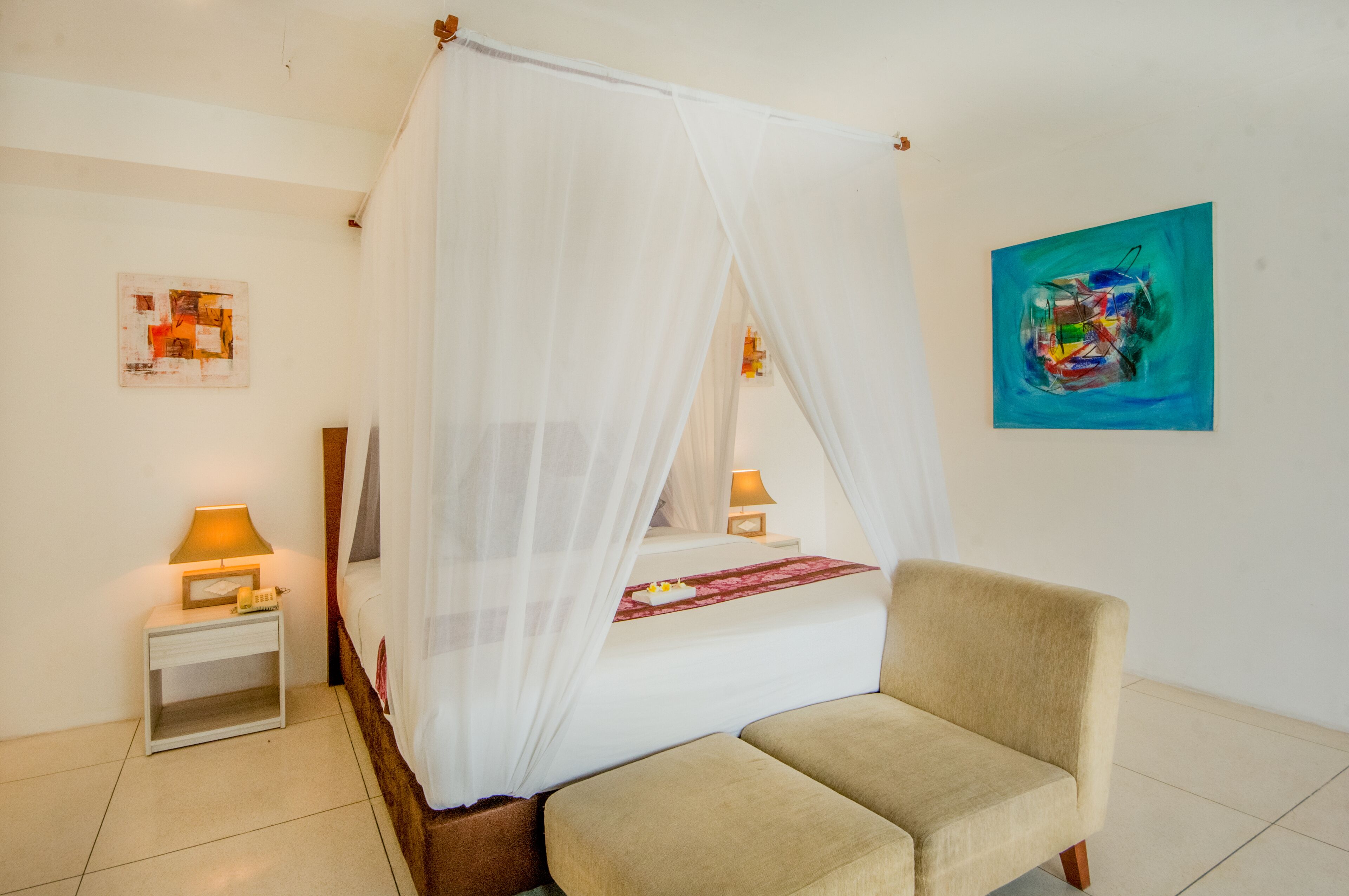 A bedroom with a four-poster bed, a bedside table with a lamp, a chair, and artwork on the walls.