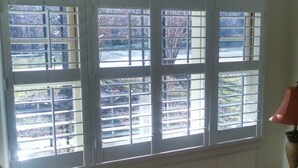 Ein Zimmer mit großem Fenster, einem Holzstuhl, einem kleinen Tisch und einer Lampe.