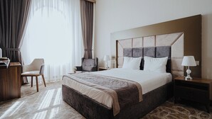 A hotel room with a large bed, a desk, a chair, a nightstand with a lamp, and a view of the outside through sheer curtains. - Volga Hotel (Baku)