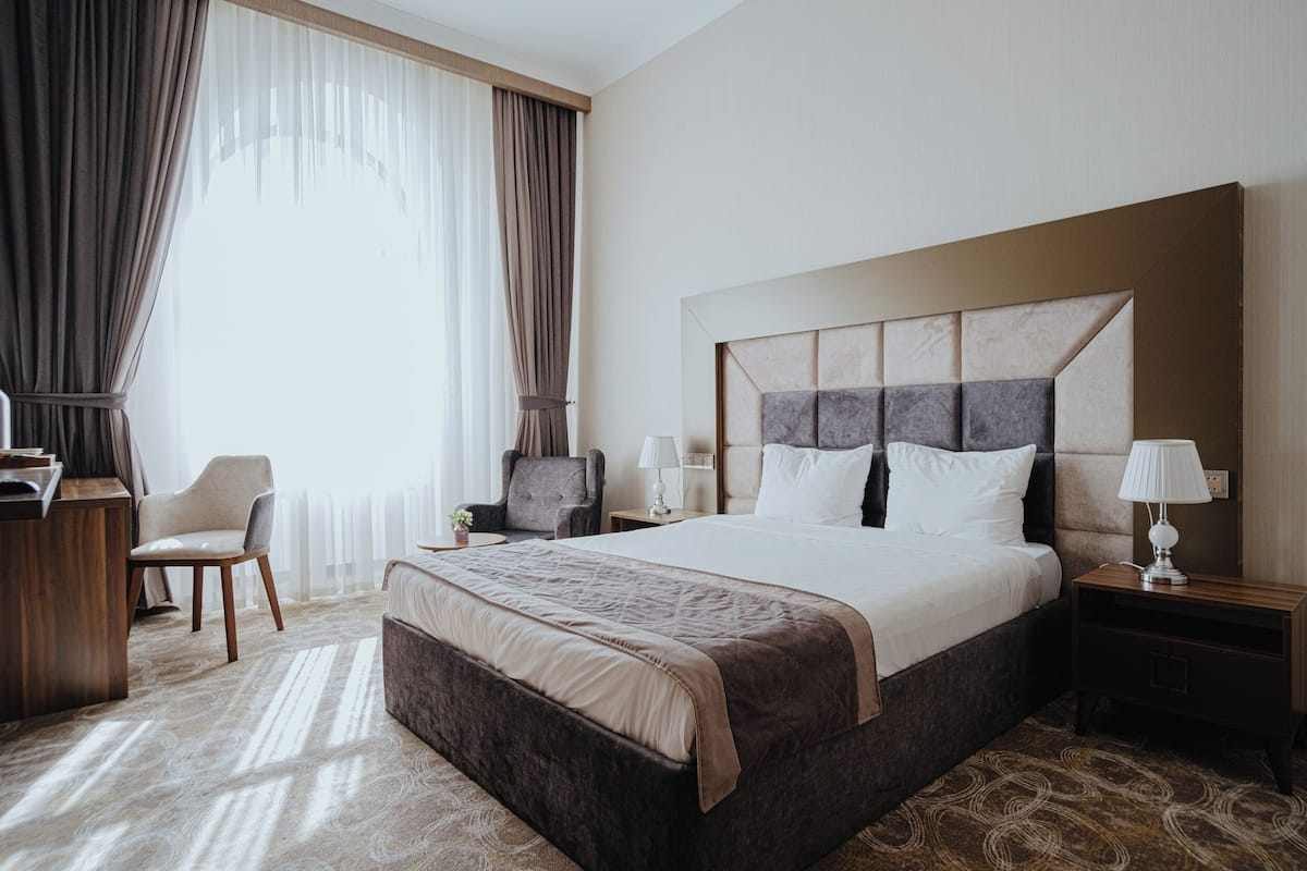 Une chambre d’hôtel avec un grand lit, un bureau, une chaise, une table de chevet avec une lampe et une vue sur l’extérieur à travers des rideaux transparents.